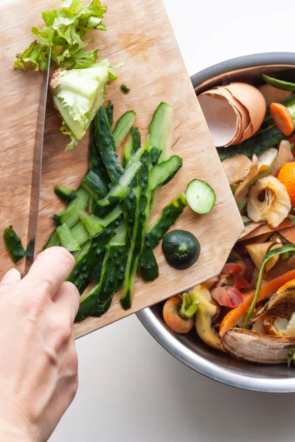 Someone dumping kitchen scraps off of a cutting board into a bucket of more scraps.