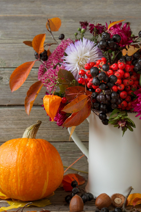 Aster flowers and other natural fall elements in a white vase next to a pumpkin. 