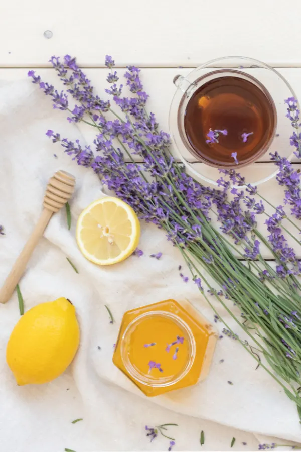 lavender blooms, lemons, and honey all lying around a cup of tea on a white tablecloth.